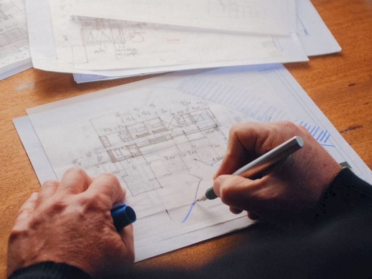 A person is drawing or reviewing architectural plans on a desk, with several papers and a pen visible.