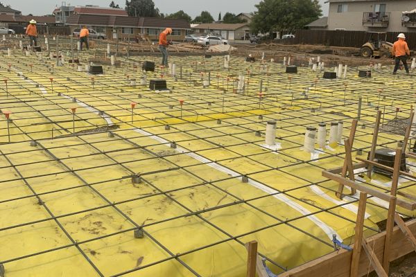 Construction site with workers, featuring a foundation with rebar grids and yellow waterproofing material, preparing for concrete pouring.