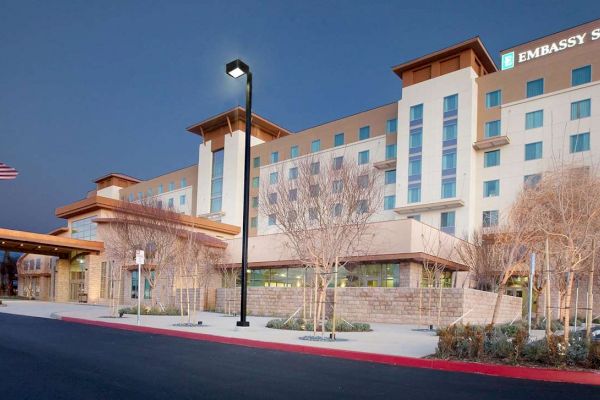 The image shows an Embassy Suites hotel building with several floors, exterior lights, an American flag, and sparse landscaping in front.