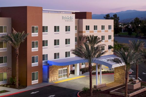 This image shows a modern four-story Fairfield Inn & Suites hotel with a front entrance, palm trees, and a parking area under a twilight sky.