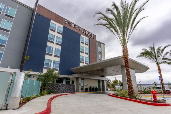 The image shows the exterior of a Springhill Suites hotel with a modern design, featuring palm trees and a driveway leading to the entrance.