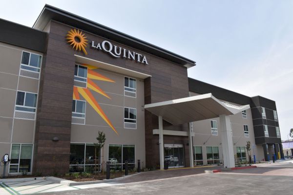 The image shows the exterior of a La Quinta hotel, featuring modern architecture with a prominent logo and a sundial design on the building.