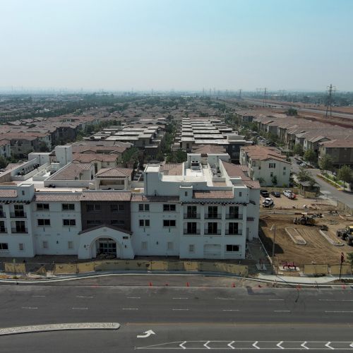 Aerial view of a residential neighborhood with houses, a large building, roads, and some open land spaces under clear skies.