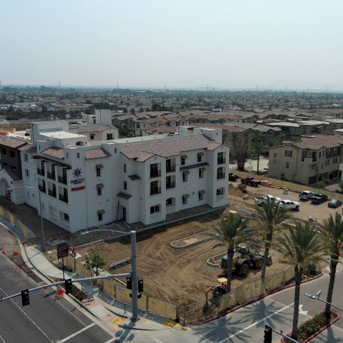 The image shows a newly constructed building next to a street, with surrounding palm trees and a residential area in the background.