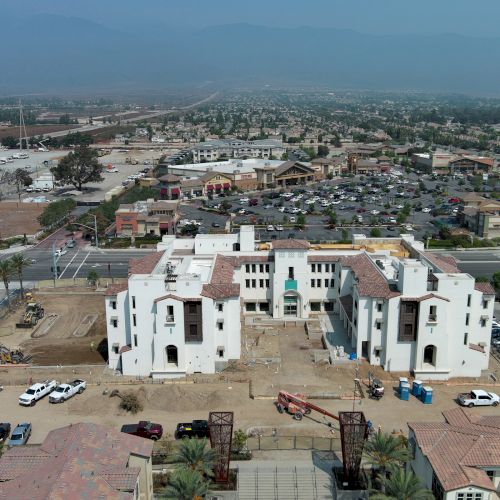 The image shows a construction site in a suburban area with buildings, parked cars, and a mountain range in the background.