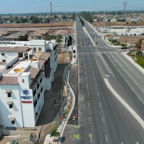 Aerial view of a road with buildings on the side, including a Hilton building, in a suburban area with clear skies.