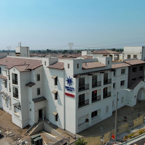 The image shows a large white, multi-story building under construction with a visible red and blue logo. There are parked vehicles and a road nearby.