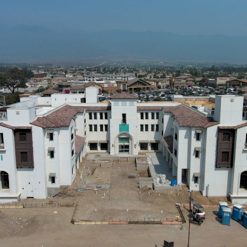 A large, white building complex under construction, surrounded by dirt and some equipment, with a cityscape and mountains in the background.