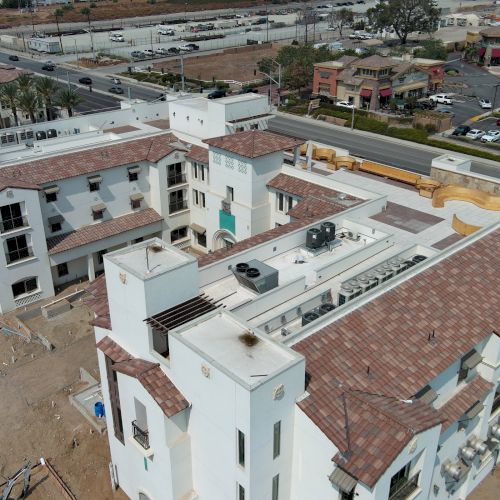 This image shows a large, white building complex with brown roofs under construction near a road, surrounded by various other buildings.