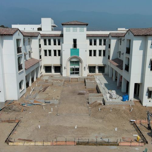 A construction site with a partially completed building featuring a central courtyard and archway, surrounded by dirt and construction materials.