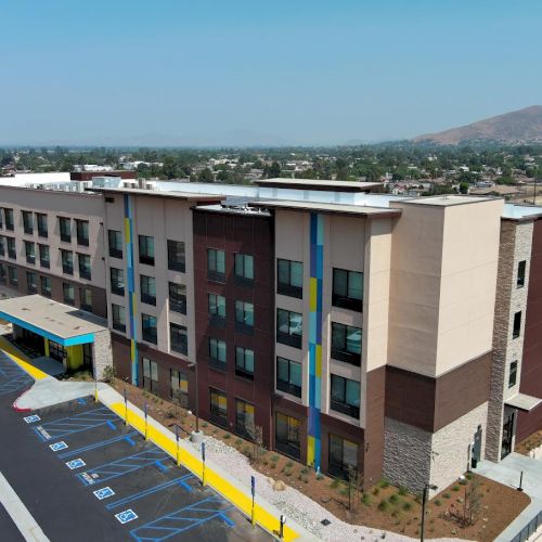 The image shows a four-story hotel building with parking spaces, including accessible parking, and a small landscaped area in an urban setting.