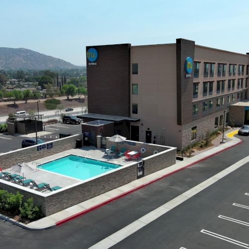 A hotel building with a swimming pool beside it, surrounded by a parking lot and landscape views in the background.