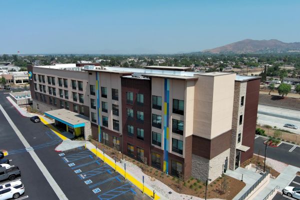 The image shows a modern, multi-story building, likely a hotel or an apartment complex, with parking spaces and a cityscape in the background.