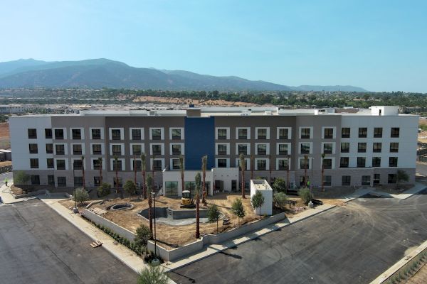 A four-story, modern building surrounded by a parking lot and landscaping, set against a mountainous backdrop under a clear blue sky.