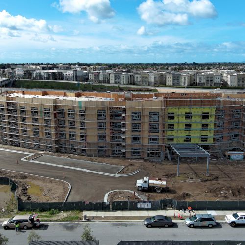 A construction site with a multi-story building in progress, surrounded by vehicles and a fenced area under a partly cloudy sky.