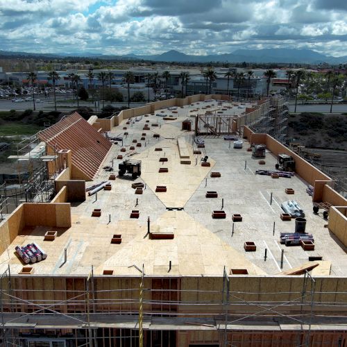 The image shows a construction site for a large building, with wooden framework and scattered equipment under a cloudy sky.