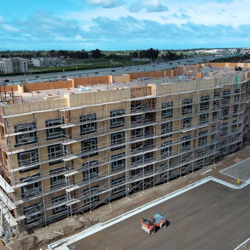 The image shows a large, multi-story building under construction, surrounded by scaffolding, and a construction vehicle on the road.