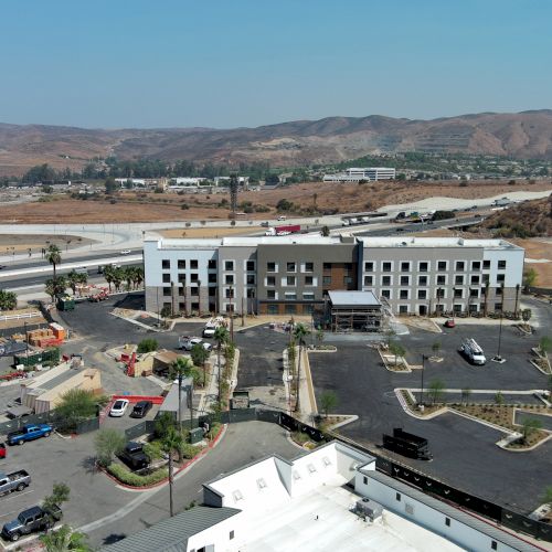 Aerial view of a multi-story building surrounded by parking lots, roads, and dry landscape with hills in the background.