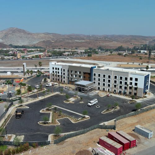 The image shows a newly constructed building with parking space, surrounded by roads and hills in the background, under a clear sky.