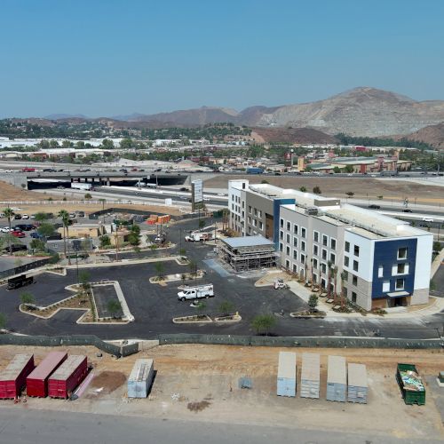 Aerial view of a developing commercial area with a building, parking lot, and construction materials, set against a mountainous backdrop.