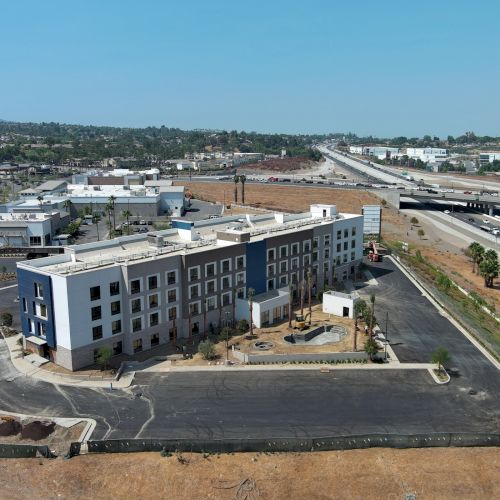 A modern, multi-story building with a large parking area is adjacent to a highway, surrounded by dry land and greenery under a clear sky.