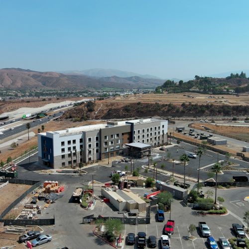 Aerial view of a building near a highway, surrounded by parking lots, cars, and hilly, dry terrain in the background.