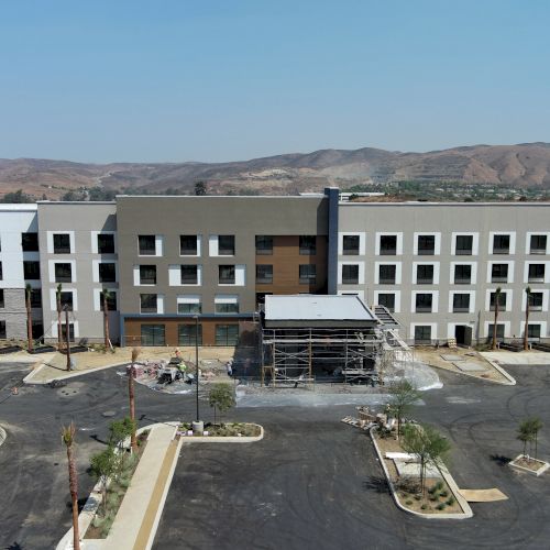 The image shows a large, modern four-story building under construction, with a parking area and some vehicles present in front.