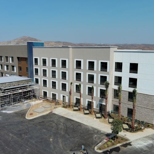 A modern multi-story building under construction with scaffolding and surrounding parking lot, situated in a semi-arid area.