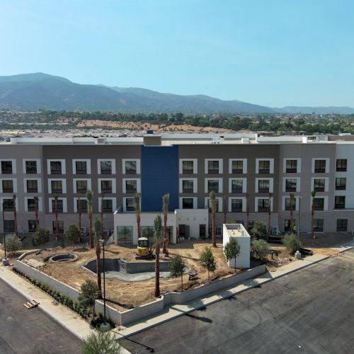 A modern four-story building with a central blue panel, surrounded by landscaped grounds and an empty parking area. Hills are visible in the background.