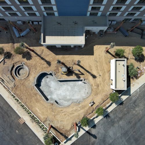 An aerial view of a construction site near a building, featuring sand, an unfinished pool, planters, and pathways.