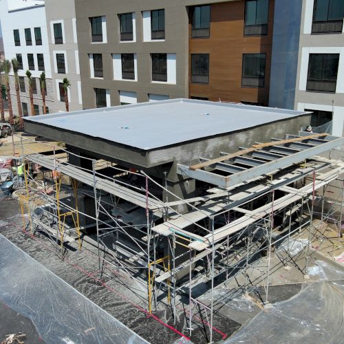 A building under construction with scaffolding and plastic sheets, adjacent to a larger completed structure.