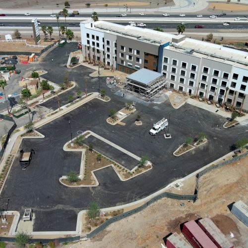 Aerial view of a large building under construction, surrounded by parking and landscaped areas, near a highway.